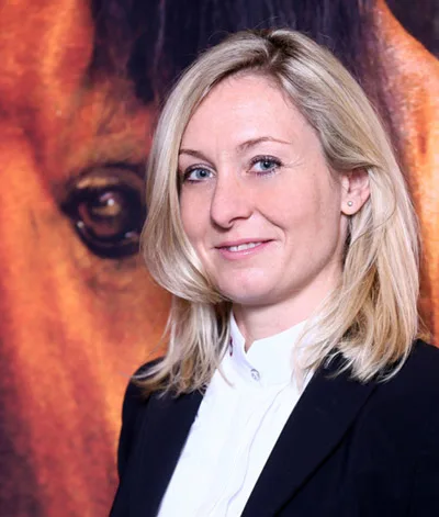 Michaela Schimak, Childéric Vertretung, im schwarzen Blazer vor einem Pferdebild