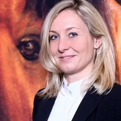 Michaela Schimak, Childéric Vertretung, im schwarzen Blazer vor einem Pferdebild